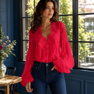 May include: A vibrant red long-sleeve blouse featuring a deep V-neckline adorned with intricate lace detailing, voluminous sleeves with ruffled cuffs, and a lightweight, flowing fabric paired with dark blue denim jeans.