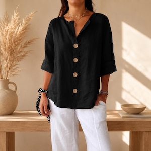 May include: A black long-sleeved shirt with a V-neckline and four large, light-colored circular buttons down the center front. The shirt features cuffed sleeves and is paired with loose-fitting white trousers.