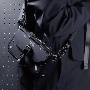 Könnte beinhalten: Eine schwarze, strukturierte Umhängetasche mit starrem, kantigem Design. Die Vorderseite zeigt das in Weiß gedruckte Wort CAUTION, begleitet von diagonalen Streifen, kleinen geometrischen Ausschnitten und einem markanten zentralen Metallverschluss.