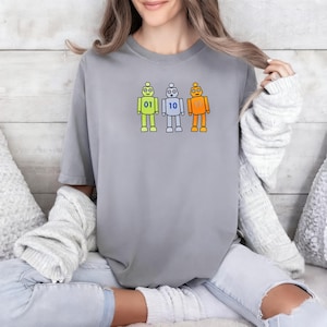 May include: A grey short-sleeved t-shirt featuring a graphic of three cartoon robots in lime green, light blue, and orange. Each robot displays a binary number on its chest: 01 on the green robot, 10 on the blue robot, and 11 on the orange robot.