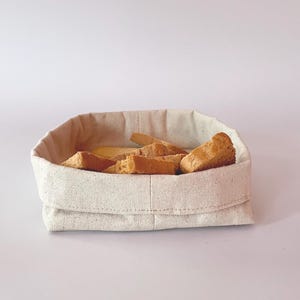 May include: A beige linen bread basket filled with several pieces of toasted bread. The basket is rectangular with a folded-over top and is sitting on a white surface. The bread pieces are golden brown and have a rough texture.