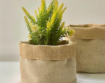 May include: Two beige fabric plant pots with a natural burlap trim. One pot contains a green and yellow plant. The pots are round and have a simple, rustic design, suitable for indoor or outdoor use.