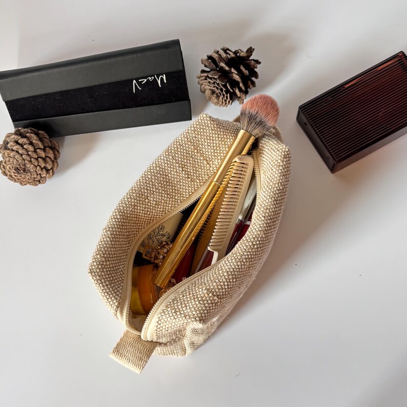May include: A beige makeup bag with a zipper, containing a makeup brush, comb, and other beauty products. A black box with the name "Maev" is visible, along with a brown compact and pine cones.
