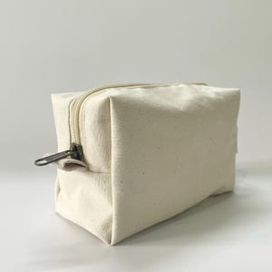 Op de afbeelding: Een rechthoekige, off-white canvas make-up tas met een beige rits en een donker metalen ritstrekker. De tas is leeg en staat op een wit oppervlak. De tas heeft een neutrale kleur en een eenvoudig ontwerp.
