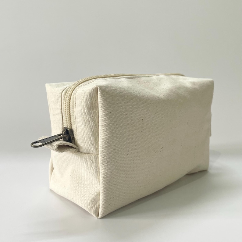 Op de afbeelding: Een rechthoekige, off-white canvas make-up tas met een beige rits en een donker metalen ritstrekker. De tas is leeg en staat op een wit oppervlak. De tas heeft een neutrale kleur en een eenvoudig ontwerp.