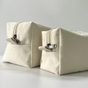 Op de afbeelding: Twee off-white canvas etuis met ritssluiting in verschillende maten. Elk etui heeft een rechthoekige vorm met een ritssluiting en een klein stoffen treklipje. De ritsen en treklipjes zijn zilverkleurig. Geschikt voor het opbergen van kleine items.