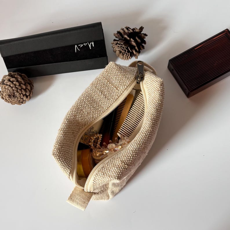 May include: A beige canvas cosmetic bag with a zipper, open to reveal combs, hair accessories, and other beauty items. A black box with the word "Maev" in silver script is visible, along with a brown rectangular box and pine cones.