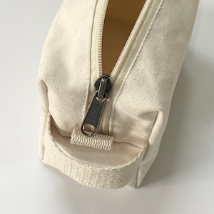 Op de afbeelding: Close-up van een cr&egrave;mekleurige canvas etui met een gedeeltelijk geopende donkere metalen YKK-rits. De etui heeft een kleine stoffen lus aan de zijkant.