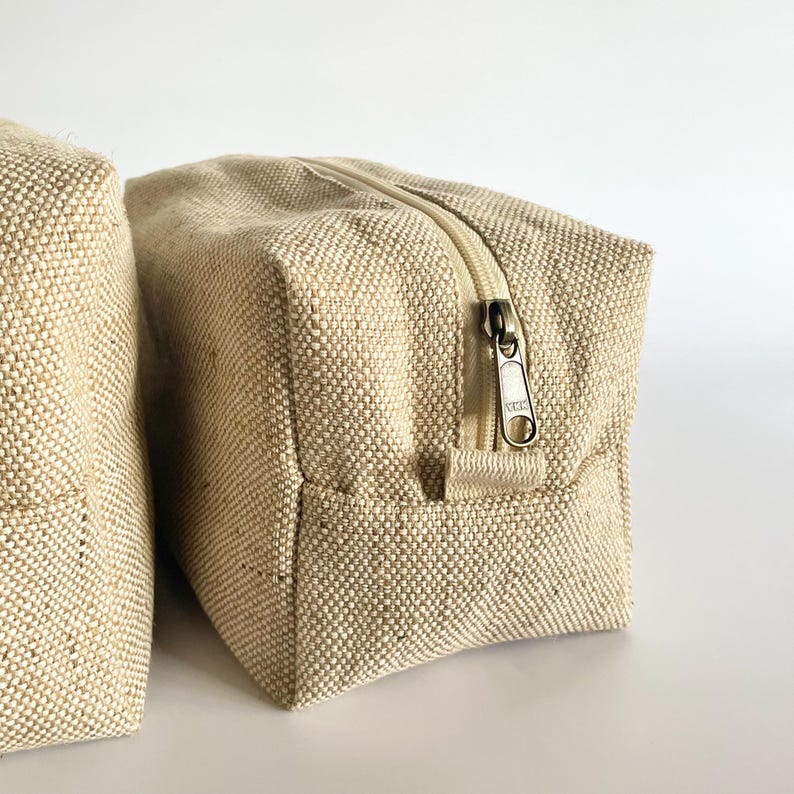 May include: Two beige canvas zippered pouches. The pouches are rectangular with a textured weave. The zippers are a light beige color with a silver-colored metal zipper pull. The pouches are likely for cosmetics or travel.