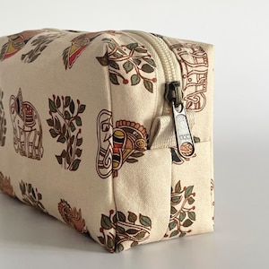 May include: A rectangular cream-colored cosmetic bag with a repeating pattern of elephants, birds, and foliage in shades of brown, red, and green. The bag features a bronze-colored zipper and a YKK zipper pull.