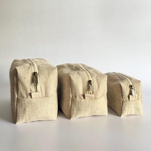 May include: Three beige canvas zippered pouches of varying sizes are arranged on a white surface. Each pouch has a rectangular shape with a zipper closure and a small fabric loop. The pouches are made of a textured, natural-looking fabric.