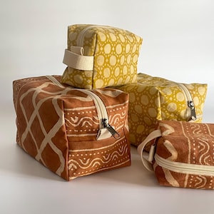 May include: Several fabric cosmetic bags in various sizes and patterns. The bags feature a repeating dot pattern in yellow and a geometric design in rust and cream. Each bag has a zipper closure and a small handle.