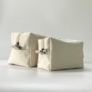 Op de afbeelding: Twee ecru canvas make-up tassen met ritssluiting. De tassen zijn rechthoekig met een vierkante basis. De ritsen zijn zilverkleurig. De tassen zijn verschillend van grootte.