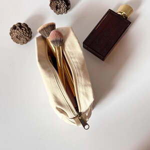 Op de afbeelding: Een beige canvas make-uptas met een gouden rits, open om make-upborstels met gouden handvatten en roze en bruine borstelharen te onthullen. Een donkerbruine parfumfles met een gouden dop staat naast de tas. Twee dennenappels zijn ook in de afbeelding.