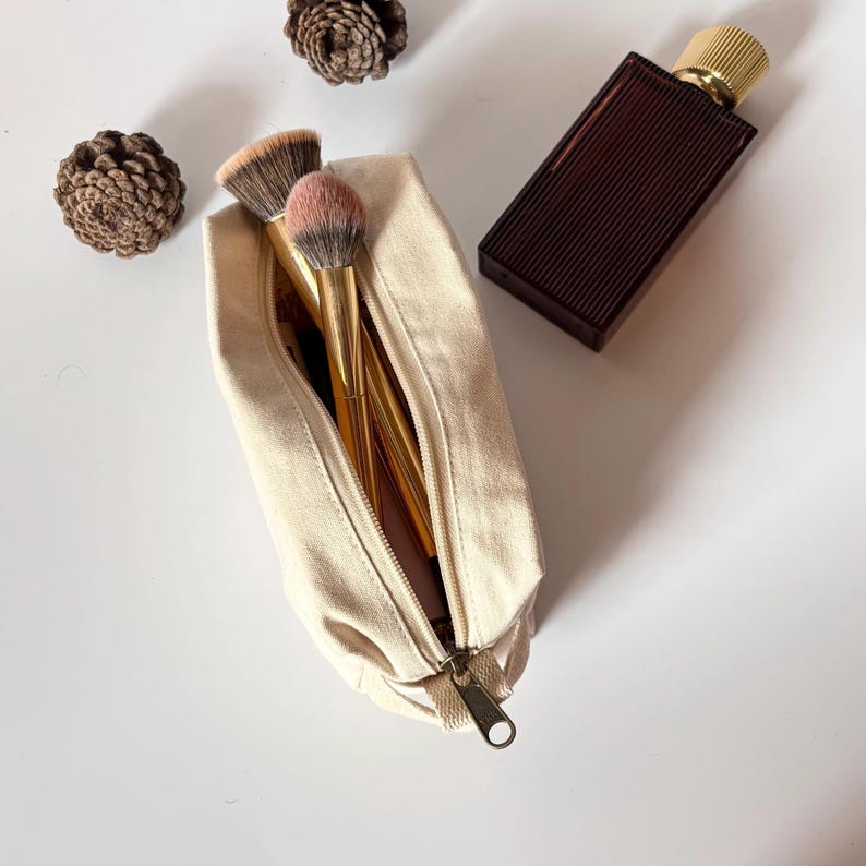 Op de afbeelding: Een beige canvas make-uptas met een gouden rits, open om make-upborstels met gouden handvatten en roze en bruine borstelharen te onthullen. Een donkerbruine parfumfles met een gouden dop staat naast de tas. Twee dennenappels zijn ook in de afbeelding.