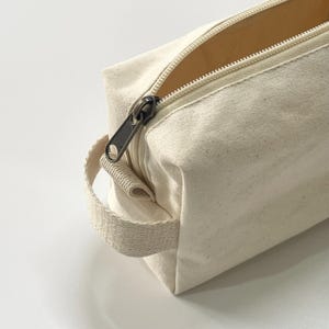Op de afbeelding: Close-up van een off-white canvas etui met ritssluiting en bijpassende handgreep. De rits is open en onthult de binnenkant. De ritstrekker en ritstanden zijn van donker metaal. De etui ligt op een witte ondergrond.