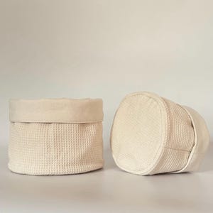 May include: Two off-white waffle-weave fabric storage baskets. One basket is upright, showing the interior and rolled-down top edge. The other basket is on its side, revealing the bottom and side construction. The baskets are made of a textured, neutral-toned fabric.
