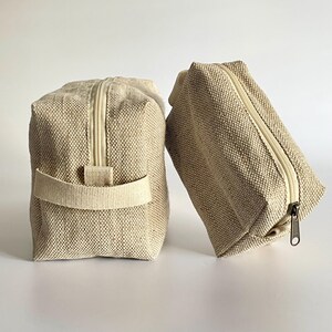 May include: Two beige canvas zippered pouches. One is upright with a handle, the other is angled. Both have a rectangular shape and a light-colored zipper. The fabric has a woven texture.