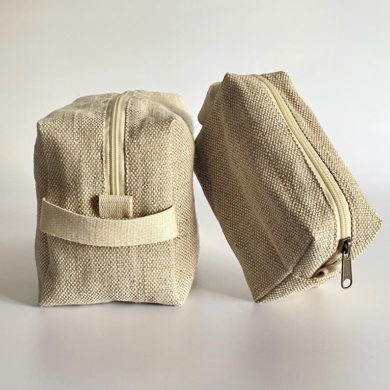 May include: Two beige canvas zippered pouches. One is upright with a handle, the other is angled. Both have a rectangular shape and a light-colored zipper. The fabric has a woven texture.