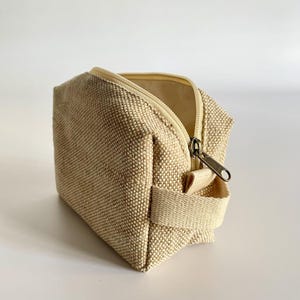 May include: Beige canvas cosmetic bag with a zipper closure and a small handle. The bag is rectangular with a textured surface and a light beige interior. The zipper is a bronze color.