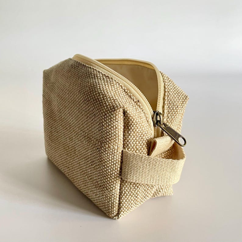 May include: Beige canvas cosmetic bag with a zipper closure and a small handle. The bag is rectangular with a textured surface and a light beige interior. The zipper is a bronze color.