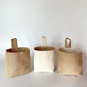 Può includere: Tre cesti portaoggetti sospesi. I cesti a sinistra e a destra sono realizzati in tessuto di iuta marrone chiaro. Il cesto centrale è in tela color crema. Ogni cesto ha un manico ad anello nella parte superiore per appenderlo.