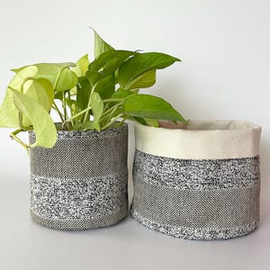 May include: Two fabric plant pots. One contains a green plant. The pots have a black and white woven pattern with a cream-colored trim. The plant has bright green and yellow leaves. The pots are cylindrical.