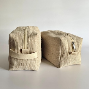 May include: Two beige rectangular toiletry bags with zipper closures. One bag has a handle on the side. The bags are made of a woven fabric and are set against a white background.