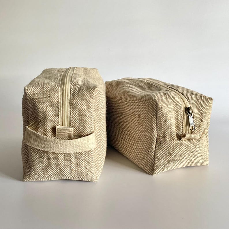 May include: Two beige rectangular toiletry bags with zipper closures. One bag has a handle on the side. The bags are made of a woven fabric and are set against a white background.