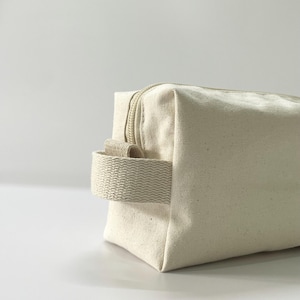 Op de afbeelding: Een close-up van een cr&egrave;mekleurige canvas toilettas. De tas heeft een rechthoekige vorm met een ritssluiting en een geweven handvat. De tas is gemaakt van een stevige, getextureerde stof.