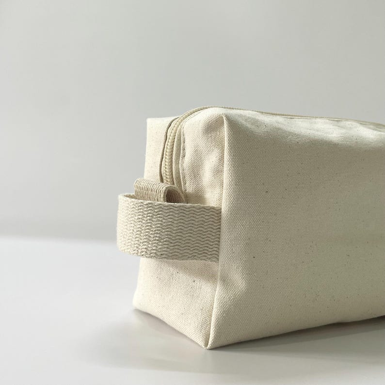 Op de afbeelding: Een close-up van een cr&egrave;mekleurige canvas toilettas. De tas heeft een rechthoekige vorm met een ritssluiting en een geweven handvat. De tas is gemaakt van een stevige, getextureerde stof.