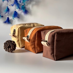 May include: Three rectangular canvas wash bags in beige, orange, and brown, each with an off-white zip and pull tab. A pinecone and blue feather dreamcatcher are in the background. These bags are ideal for travel or everyday use.