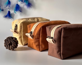 Op de afbeelding: Drie rechthoekige canvas toilettassen in beige, oranje en bruin, elk met een contrasterende crèmekleurige ritssluiting en treklipje. Een dennenappel en een dromenvanger met blauwe veren staan op de achtergrond.