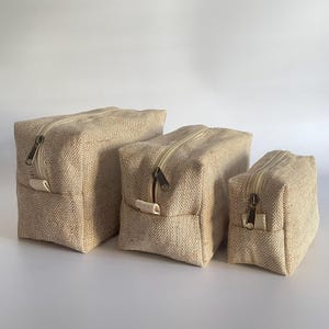 May include: Three beige canvas zippered pouches of varying sizes. Each pouch has a rectangular shape with a beige zipper and a small fabric loop on one side. The pouches are made of a textured, woven material.