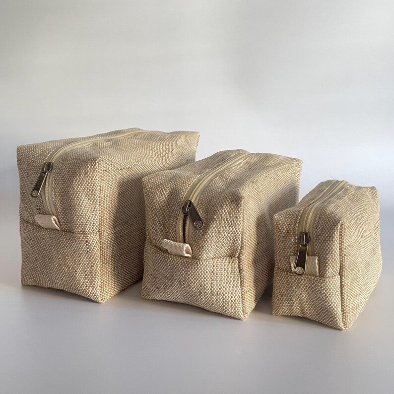 May include: Three beige canvas zippered pouches of varying sizes. Each pouch has a rectangular shape with a beige zipper and a small fabric loop on one side. The pouches are made of a textured, woven material.