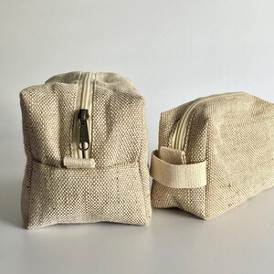 May include: Two beige canvas zippered pouches. The pouches are rectangular with a textured weave. One pouch has a small handle on the side. The zippers are a light beige color with a dark metal zipper pull. The pouches are sitting on a white surface.