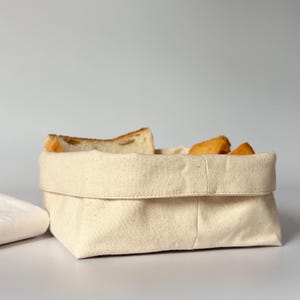 May include: A rectangular, off-white canvas bread basket with a folded-over top, filled with slices of toasted bread. The basket sits on a white surface, suggesting a kitchen or dining setting. The neutral tones create a clean, minimalist aesthetic.