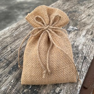 Puede incluir: Una pequeña bolsa de arpillera beige con cierre de cordón. La bolsa está atada con un lazo de cuerda natural en la parte superior. La bolsa está sobre una superficie de madera envejecida.