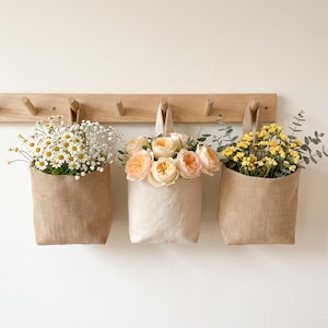 May include: Three hanging fabric pouches suspended from a light wood wall rack. The left and right pouches are made of tan hessian, while the centre pouch is off-white canvas. They contain white daisies, peach roses, and yellow flowers.
