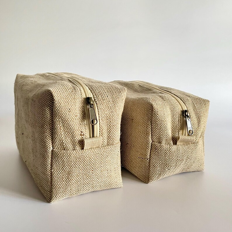 May include: Two beige rectangular zippered pouches. The pouches are made of a textured fabric and have a rectangular shape. The zippers are a light beige color. The pouches are likely for storing toiletries or other small items.