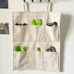 May include: A beige canvas hanging organiser with multiple pockets, holding various kitchen utensils. The organiser is suspended from a wall hook. Utensils include green spatulas, a peeler, and scissors.