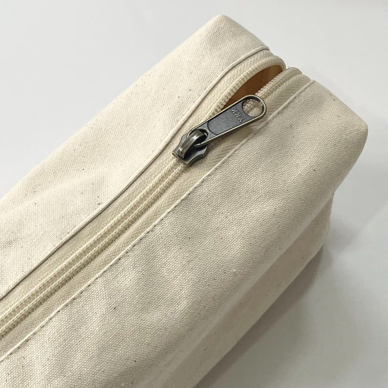 Op de afbeelding: Close-up van een cr&egrave;mekleurige canvas etui met een metalen ritssluiting. De ritssluiting is gedeeltelijk geopend en onthult de binnenkant. Het canvas materiaal heeft een licht getextureerde uitstraling.