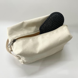 Op de afbeelding: Een cr&egrave;mekleurige canvas cosmetische tas met een ritssluiting en een zwarte borstel erin. De tas heeft een klein handvat aan de zijkant. De borstel is zwart met een getextureerd oppervlak.