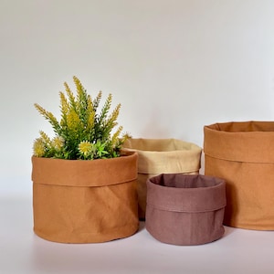 Op de afbeelding: Vier stoffen plantenpotten in bruin- en beigetinten. Eén pot bevat een plant met geel en groen blad. De potten variëren in grootte en zijn gerangschikt op een wit oppervlak, wat een minimalistisch ontwerp laat zien.