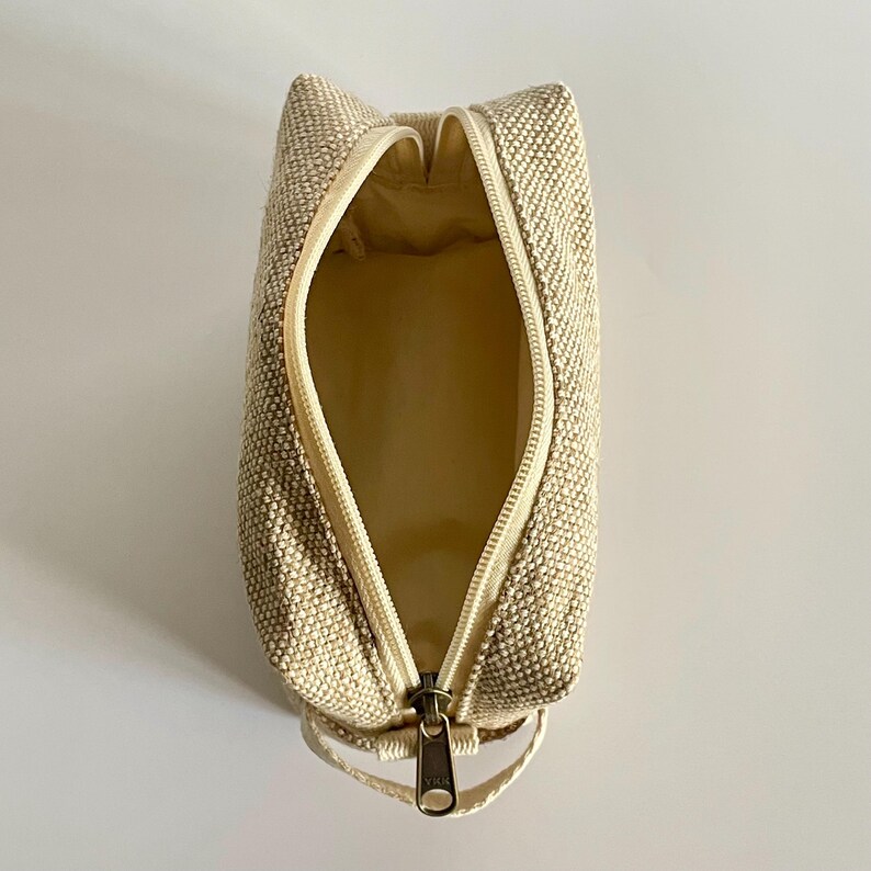 May include: Open, beige-colored rectangular cosmetic bag with a zipper closure. The bag is made of a textured fabric and has a matching beige interior. The zipper is open, revealing the inside of the bag.