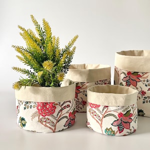 May include: Four fabric storage baskets in different sizes. The baskets have a cream-coloured interior and a floral pattern with red, pink, green, and brown accents on a cream background. One basket contains a green and yellow plant.