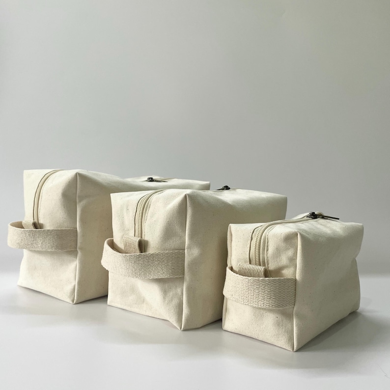 Op de afbeelding: Drie off-white canvas ritszakjes in verschillende maten. Elk heeft een bijpassende stoffen handgreep en een donkere rits. De zakjes zijn in een rij gerangschikt en tonen hun afmetingen.