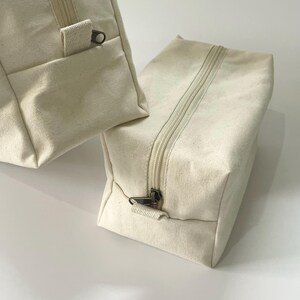 Op de afbeelding: Twee off-white canvas tassen met ritssluiting. E&eacute;n tas heeft een klein vakje aan de voorkant. De ritsen en ritstrekkers zijn donker metaalkleurig. De tassen zijn rechthoekig en lijken leeg te zijn.
