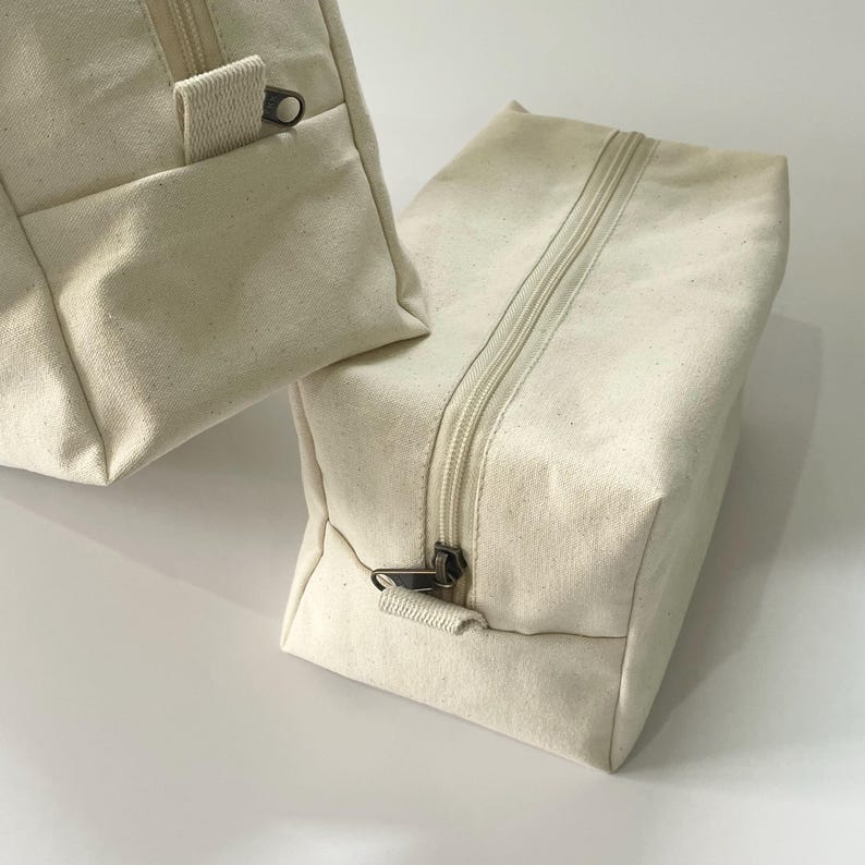 Op de afbeelding: Twee off-white canvas tassen met ritssluiting. E&eacute;n tas heeft een klein vakje aan de voorkant. De ritsen en ritstrekkers zijn donker metaalkleurig. De tassen zijn rechthoekig en lijken leeg te zijn.