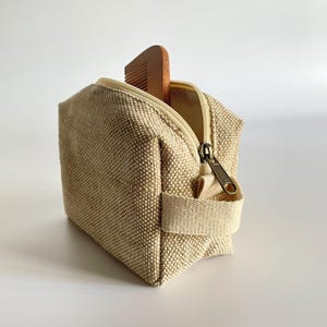 May include: A small, beige canvas toiletry bag with a zipper and a handle. A wooden comb is partially visible inside the open bag. The bag has a textured appearance and a neutral color palette.
